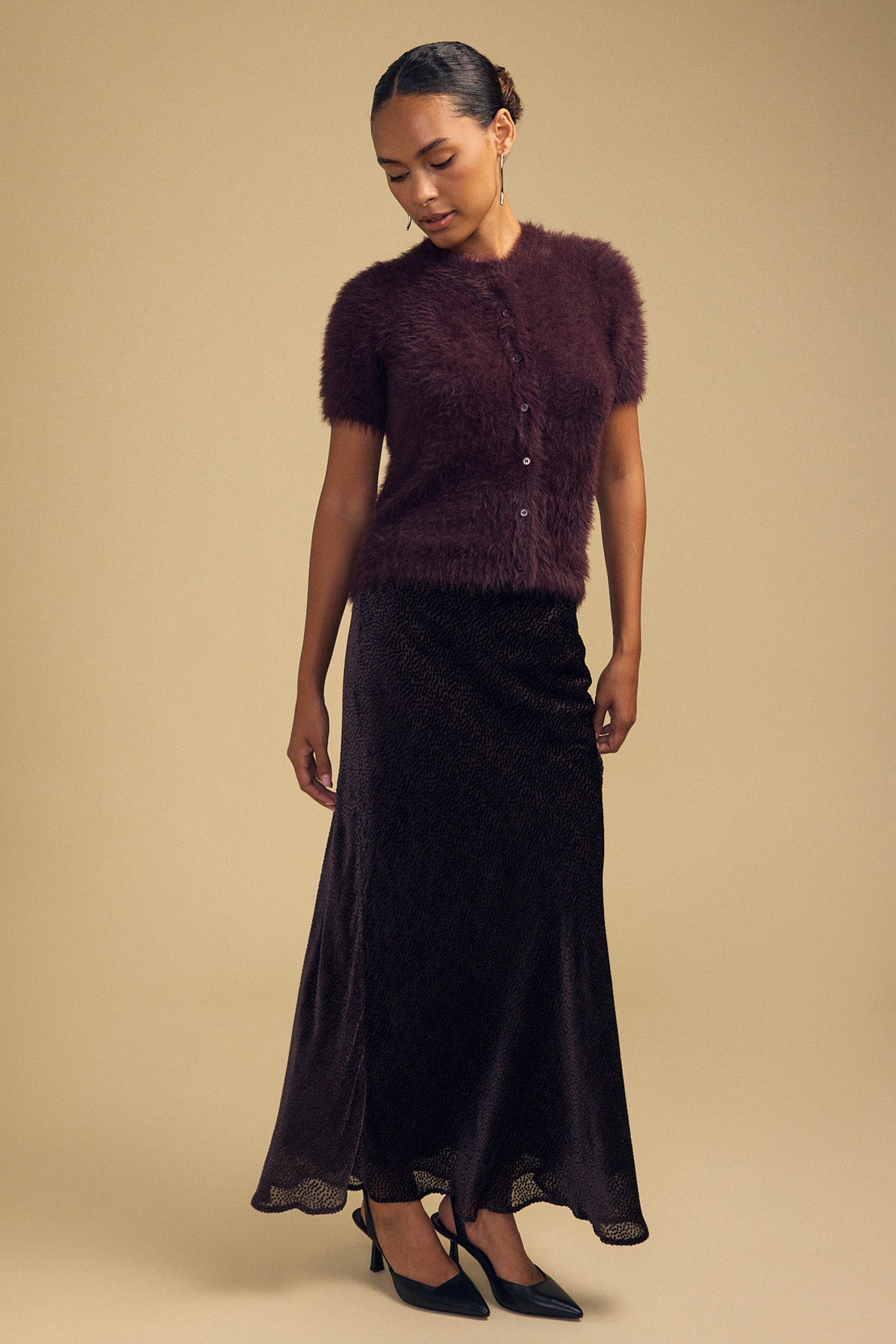 A model wears a dark purple GABBY FEATHER YARN CARDIGAN, a soft, airy, short-sleeve button-down, paired with a long dark patterned skirt and black pointed-toe heels.