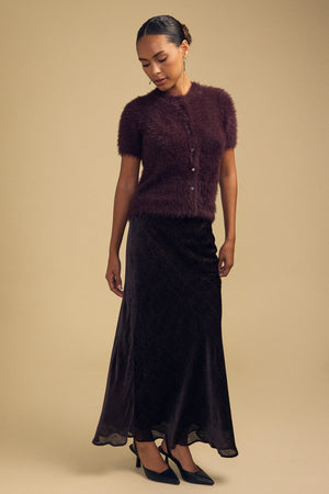 A model wears a dark purple GABBY FEATHER YARN CARDIGAN, a soft, airy, short-sleeve button-down, paired with a long dark patterned skirt and black pointed-toe heels.