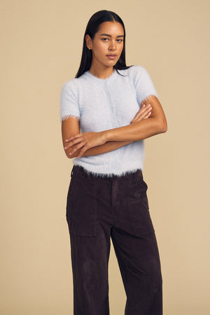 A woman wearing a pale blue, soft airy short-sleeved GABBY FEATHER YARN CARDIGAN by Velvet by Graham & Spencer paired with dark brown trousers.