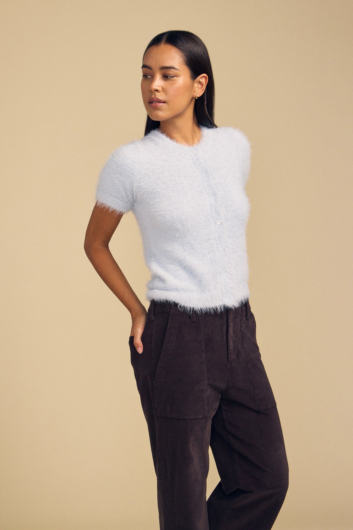 A woman wears a light blue, short-sleeve fuzzy knit GABBY FEATHER YARN CARDIGAN buttoned up with dark trousers.