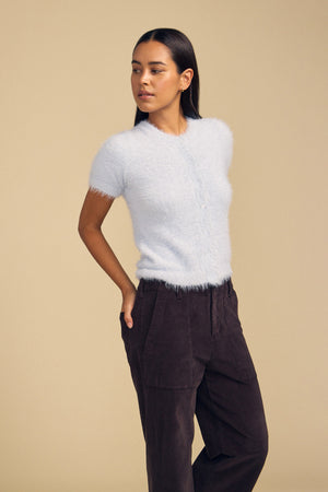 A woman wears a light blue, short-sleeve fuzzy knit GABBY FEATHER YARN CARDIGAN buttoned up with dark trousers.