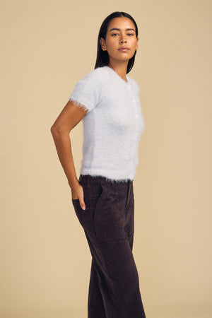 A person wearing a soft, airy light blue GABBY FEATHER YARN CARDIGAN with short sleeves and dark trousers.