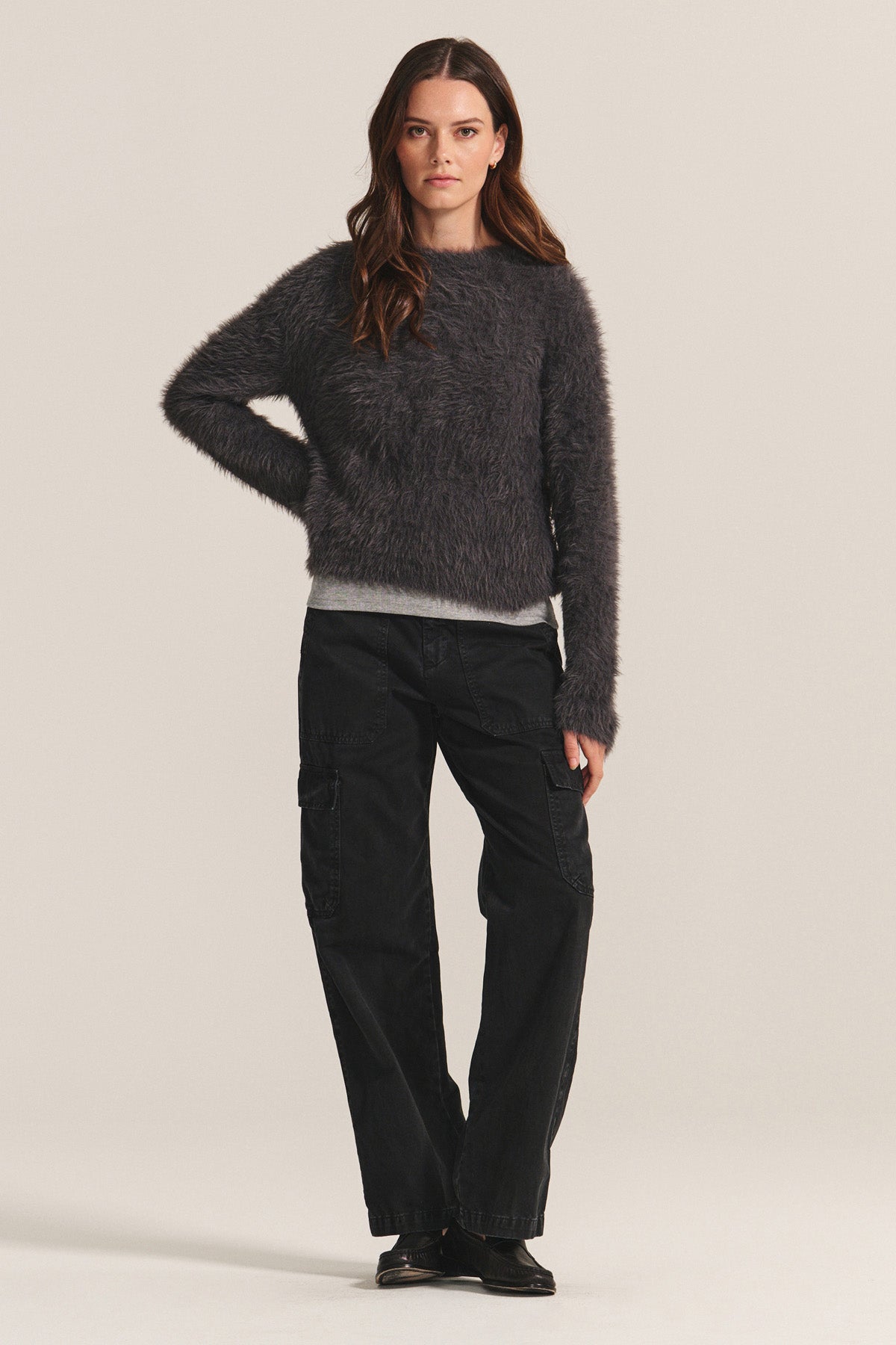 A woman faces forward, wearing the PENNY SWEATER by Velvet by Graham & Spencer—a lightweight feather yarn dark gray crewneck—paired with black cargo pants and shoes, set against a plain light background.