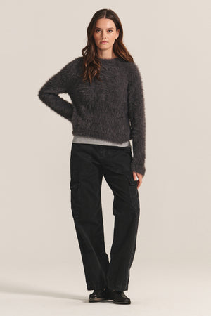 A woman faces forward, wearing the PENNY SWEATER by Velvet by Graham & Spencer—a lightweight feather yarn dark gray crewneck—paired with black cargo pants and shoes, set against a plain light background.