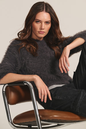 A woman with long brown hair wears the PENNY SWEATER by Velvet by Graham & Spencer—a dark, fuzzy crew neck—paired with black pants as she sits on a brown chair, looking at the camera with a neutral expression.