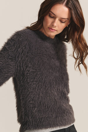 A woman looks down, her loose waves framing her face as she wears the Velvet by Graham & Spencer PENNY SWEATER, a long-sleeved, dark gray fuzzy piece with a classic crew neckline.