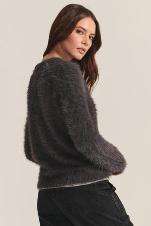 A woman models the PENNY SWEATER by Velvet by Graham & Spencer, a long-sleeve, dark gray feather yarn knit, paired with black jeans as she poses sideways against a plain background.