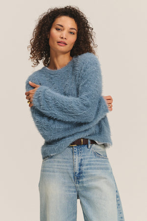 Wearing the PENNY SWEATER by Velvet by Graham & Spencer—a soft, fuzzy blue crewneck—paired with light blue jeans, this person stands against a plain backdrop, radiating versatile comfort.