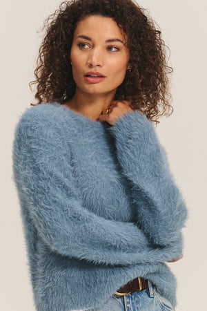 A woman in Velvet by Graham & Spencer’s PENNY SWEATER—a soft blue, lightweight feather yarn crewneck—stands with arms crossed in jeans against a plain light background, highlighting the sweater’s versatile comfort.