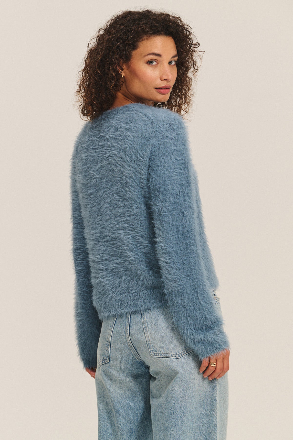 Someone with curly hair wears the PENNY SWEATER by Velvet by Graham & Spencer, a lightweight feather yarn crewneck, paired with light blue jeans. They stand against a plain background, looking over their shoulder.-40248224579777