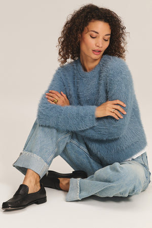A woman with curly hair sits on the floor with arms crossed, wearing the PENNY SWEATER by Velvet by Graham & Spencer—a blue fuzzy piece crafted from lightweight feather yarn—paired with light blue jeans and black loafers against a neutral background.