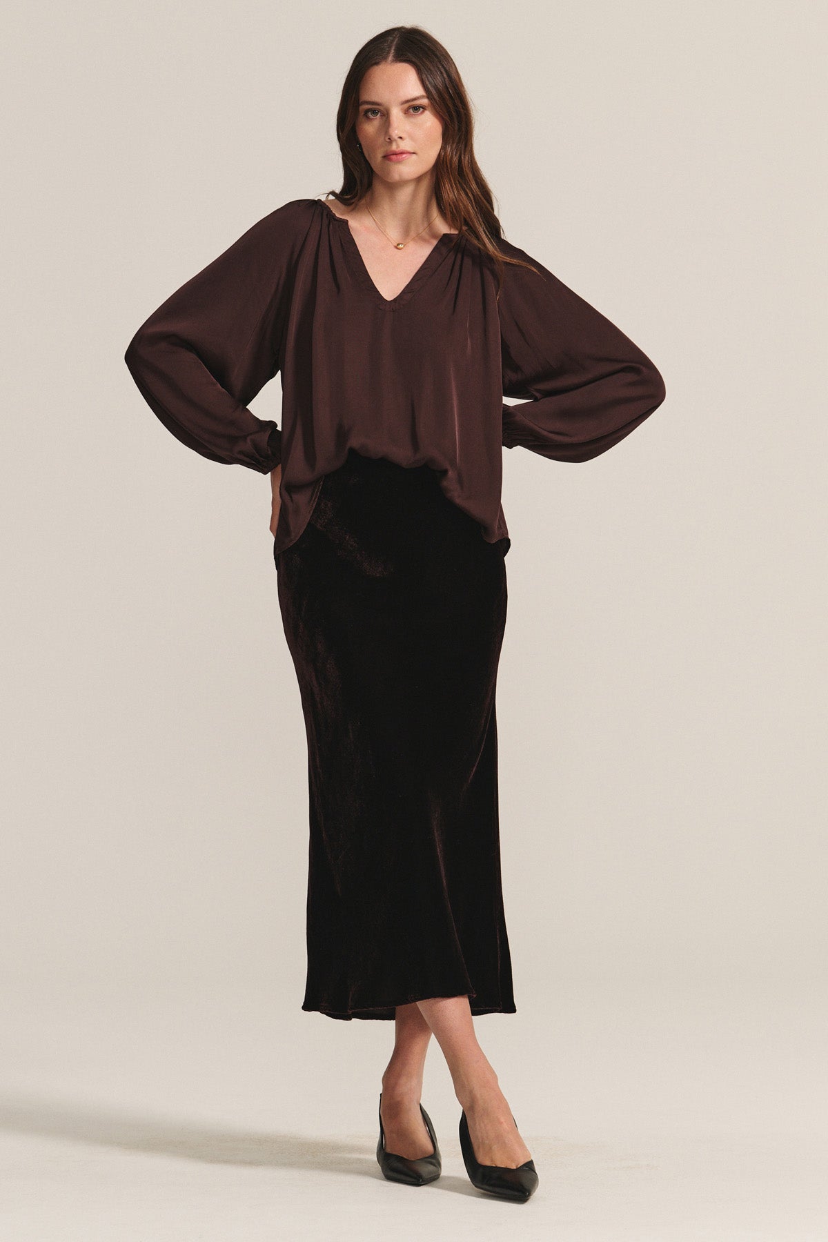 A woman stands against a plain background, wearing a loose brown blouse and the FELICITY SKIRT by Velvet by Graham & Spencer—a black silk velvet skirt with an elastic waistband—paired with black heels, arms akimbo and a neutral expression.-40263648641217