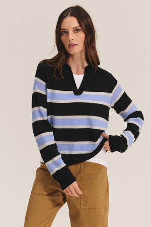 A person wearing the Velvet by Graham & Spencer EVERLAKE SWEATER POLO, featuring black, blue, and white stripes, over a white shirt with tan pants, stands against a plain background.