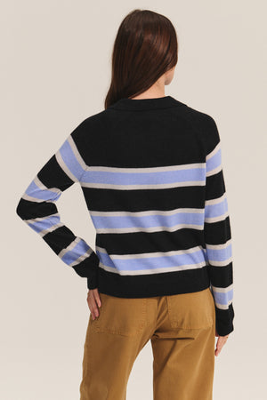 A person with long brown hair, wearing the EVERLAKE SWEATER POLO by Velvet by Graham & Spencer—black with blue and white stripes—and tan pants stands with their back to the camera.