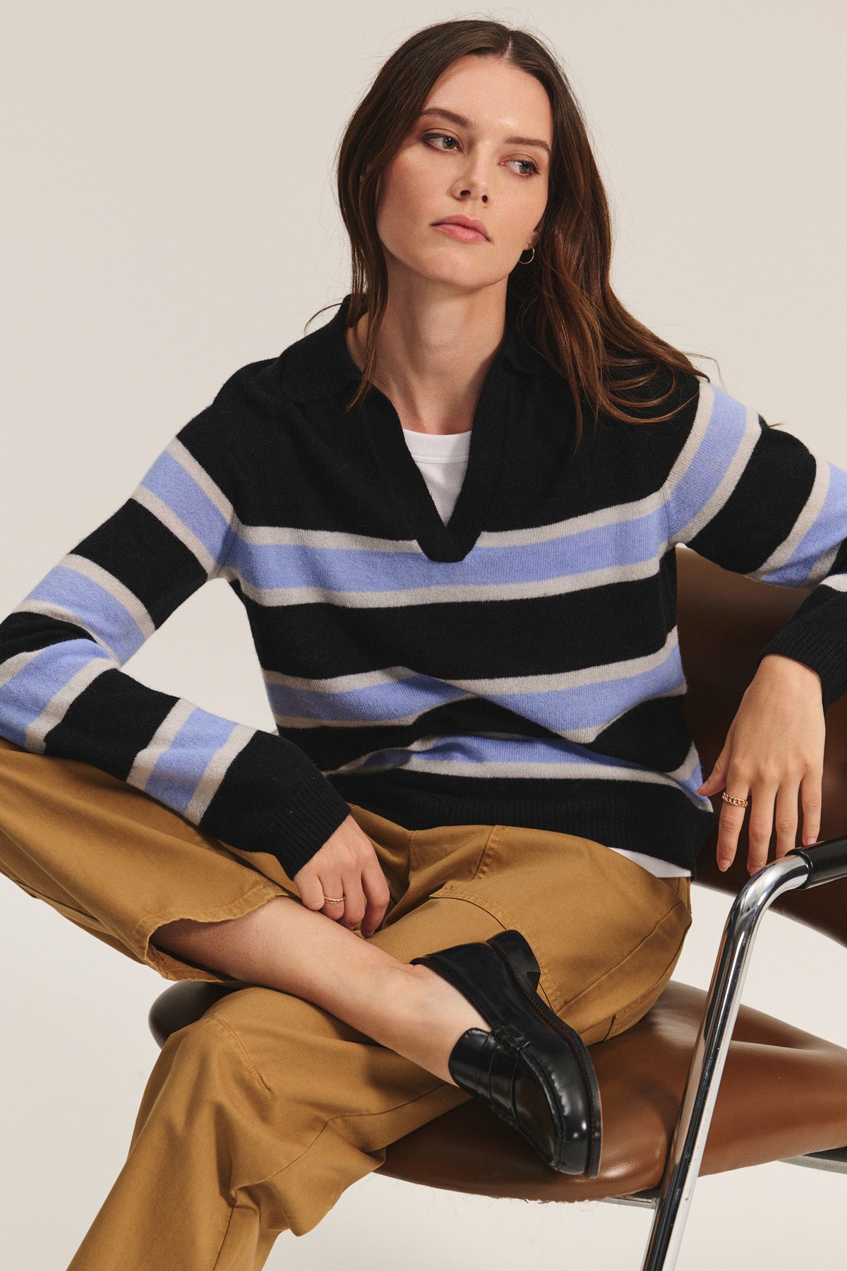 A woman sits on a brown chair in the EVERLAKE SWEATER POLO by Velvet by Graham & Spencer—a black, white, and blue striped alpaca-blend V-neck sweater—paired with tan pants and black loafers, her arms relaxed and legs crossed.