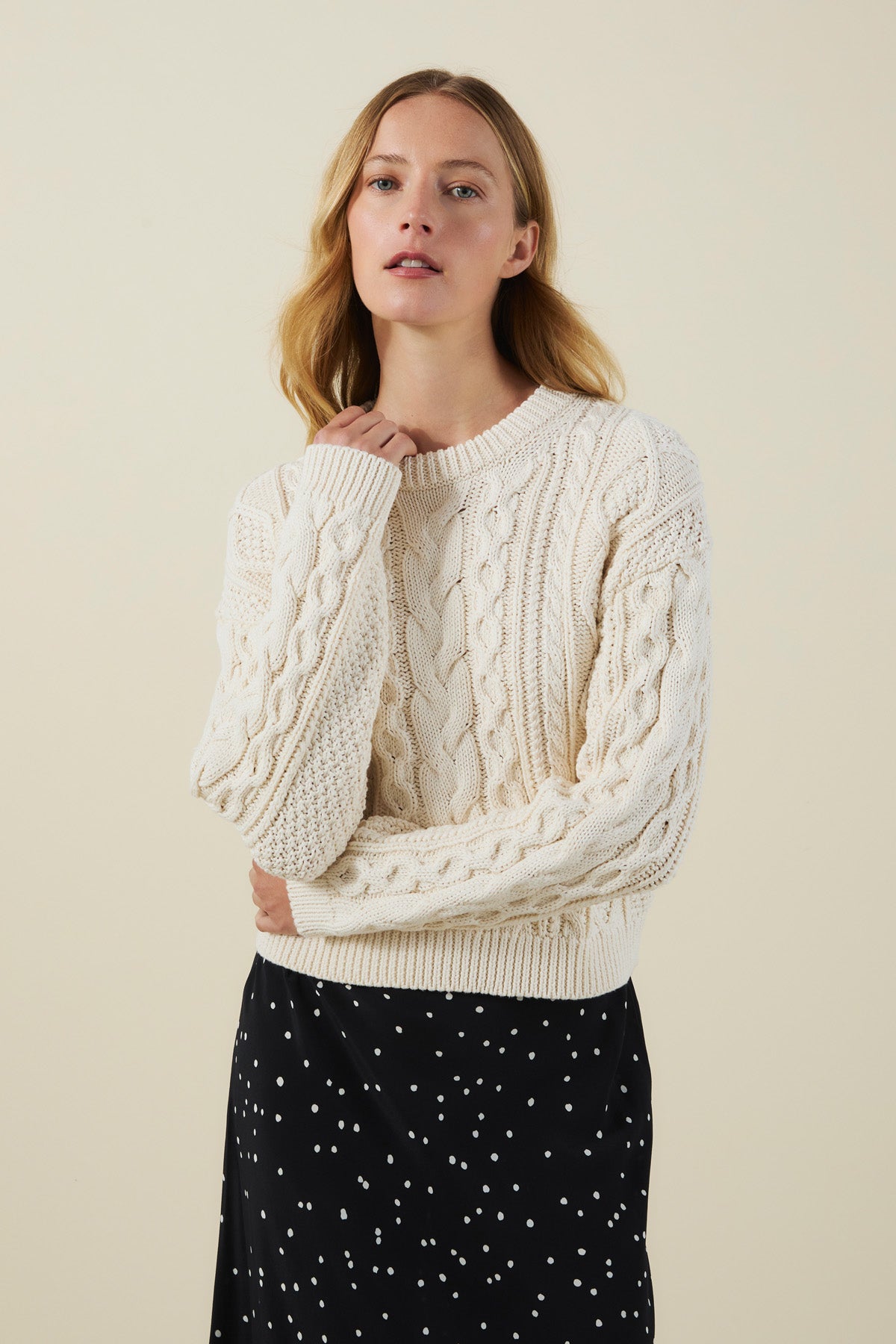 A woman wearing the EVA SWEATER by Velvet by Graham & Spencer, a cream-colored cable knit piece, and a black skirt with white dots poses against a plain beige background.-41826809839809