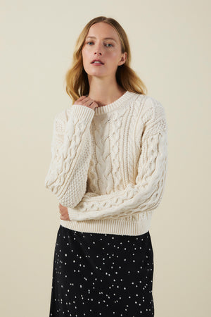 A woman wearing the EVA SWEATER by Velvet by Graham & Spencer, a cream-colored cable knit piece, and a black skirt with white dots poses against a plain beige background.