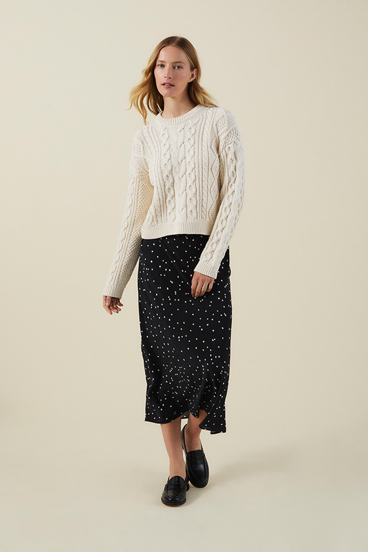 A person wearing a cream cable-knit sweater, the CADENCE SILK SKIRT by Velvet by Graham & Spencer—a black skirt with white polka dots—and black loafers stands against a plain light background.-41970981535937