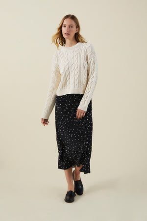 A person wearing a cream cable-knit sweater, the CADENCE SILK SKIRT by Velvet by Graham & Spencer—a black skirt with white polka dots—and black loafers stands against a plain light background.