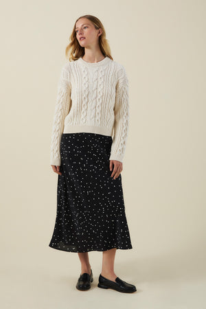A woman stands against a plain beige background wearing a cream cable-knit sweater, black loafers, and the CADENCE SILK SKIRT by Velvet by Graham & Spencer—a classic A-line skirt featuring a black polka dot print.