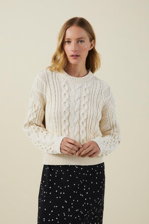Model wearing the EVA SWEATER by Velvet by Graham & Spencer in cream, paired with a black skirt with white polka dots, standing against a plain light background.