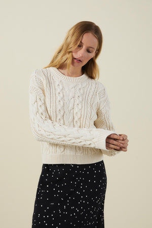 A woman models the EVA SWEATER by Velvet by Graham & Spencer, a cream cable knit design, paired with a black skirt featuring white polka dots against a plain background.