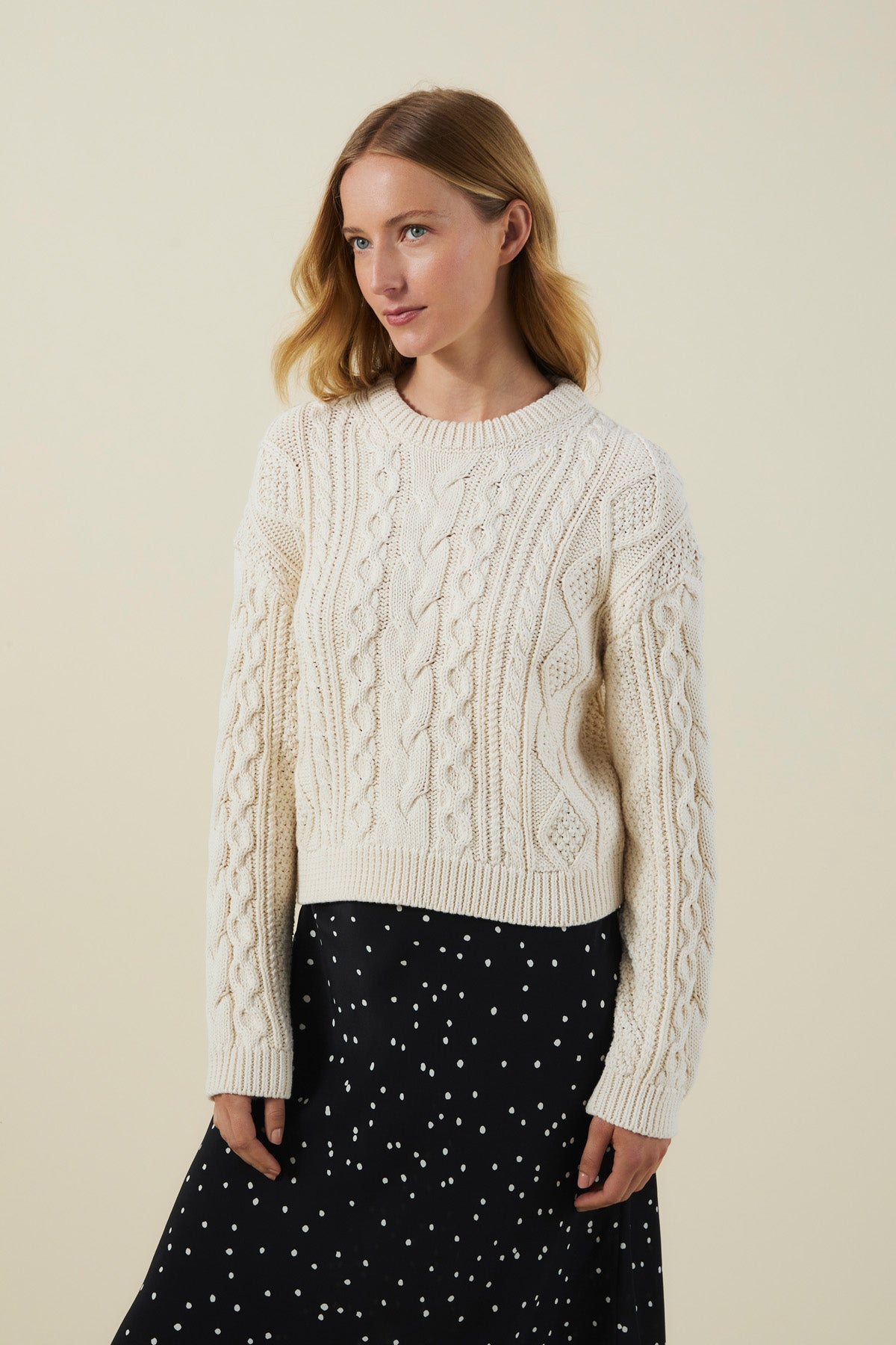 A woman with light brown hair wears the EVA SWEATER by Velvet by Graham & Spencer, a cream cable knit design, paired with a black skirt with white dots, standing against a plain beige background.