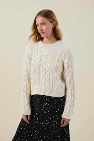 A woman with light brown hair wears the EVA SWEATER by Velvet by Graham & Spencer, a cream cable knit design, paired with a black skirt with white dots, standing against a plain beige background.