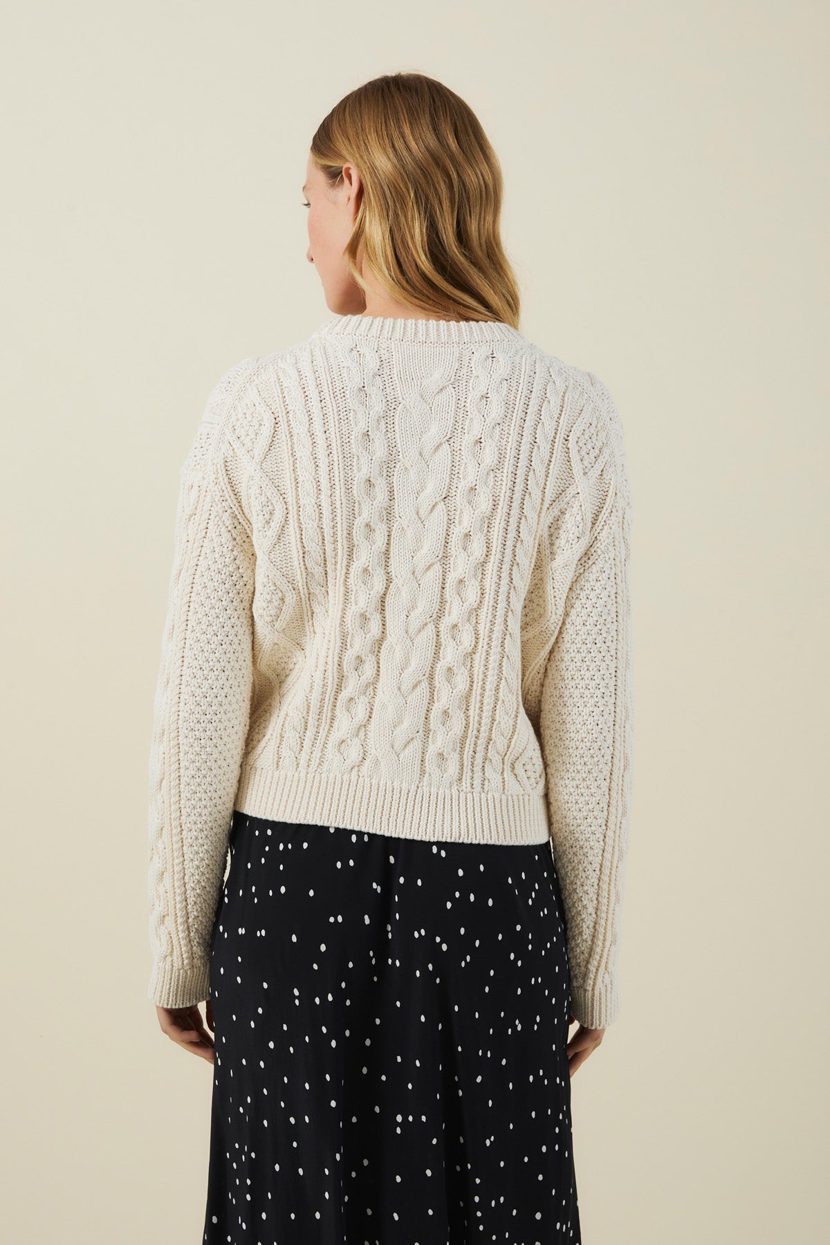 A person with wavy, light brown hair wears the EVA SWEATER by Velvet by Graham & Spencer—a cream cable-knit design—paired with a black skirt featuring white dots, facing away from the camera against a plain background.-41826809708737