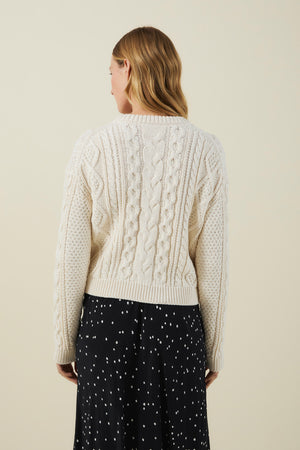 A person with wavy, light brown hair wears the EVA SWEATER by Velvet by Graham & Spencer—a cream cable-knit design—paired with a black skirt featuring white dots, facing away from the camera against a plain background.