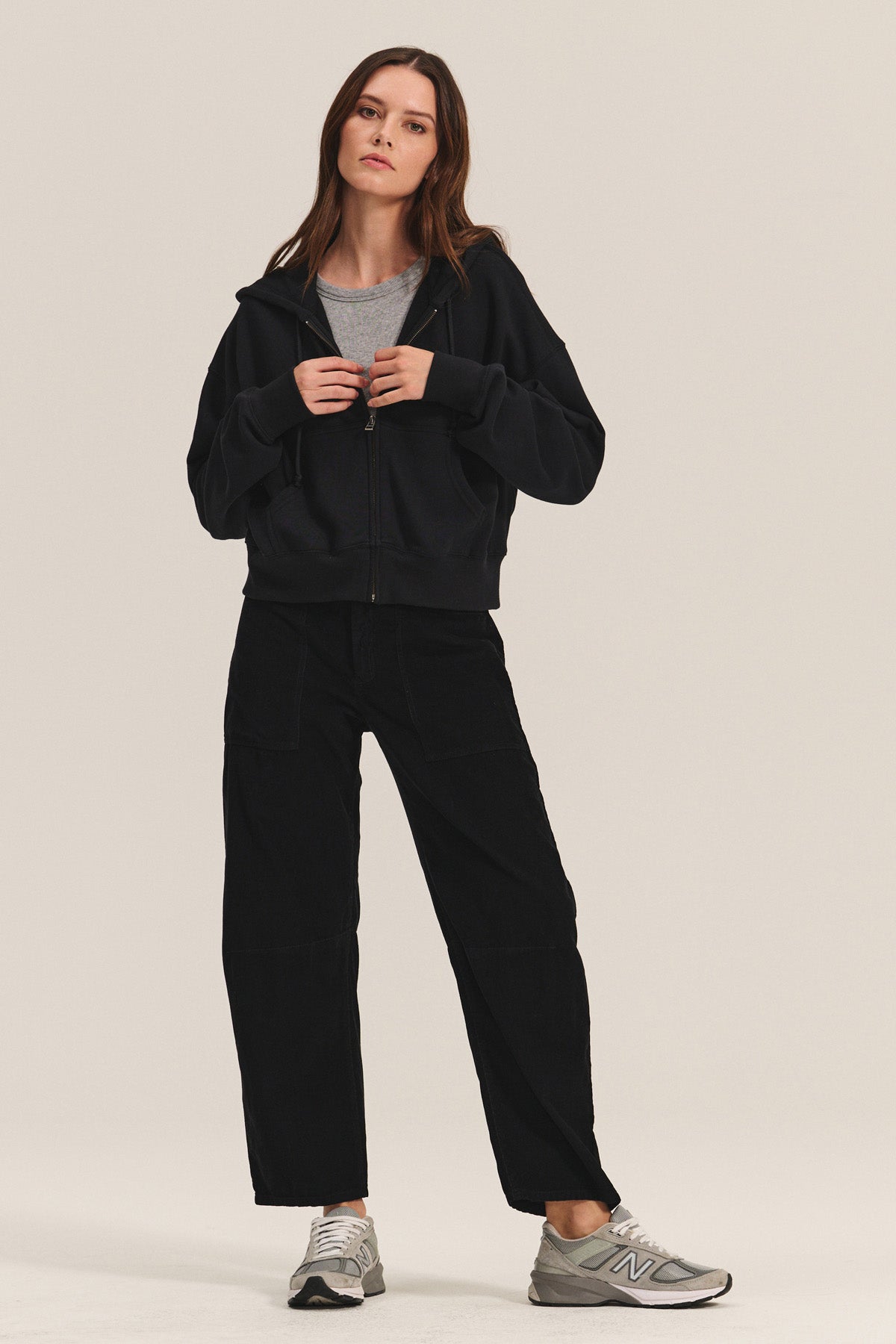 A woman stands in a neutral pose wearing the Velvet by Graham & Spencer HIGHLAND HOODIE, a black fleece full zip hoodie with drawstring hood, paired with a gray t-shirt, black pants, and gray New Balance sneakers against a plain background.-40293783339201