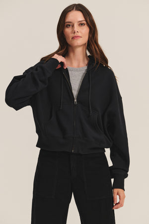 A woman stands against a plain background, wearing Velvet by Graham & Spencer’s HIGHLAND HOODIE in black over a gray t-shirt, with one hand touching the hoodie’s collar.