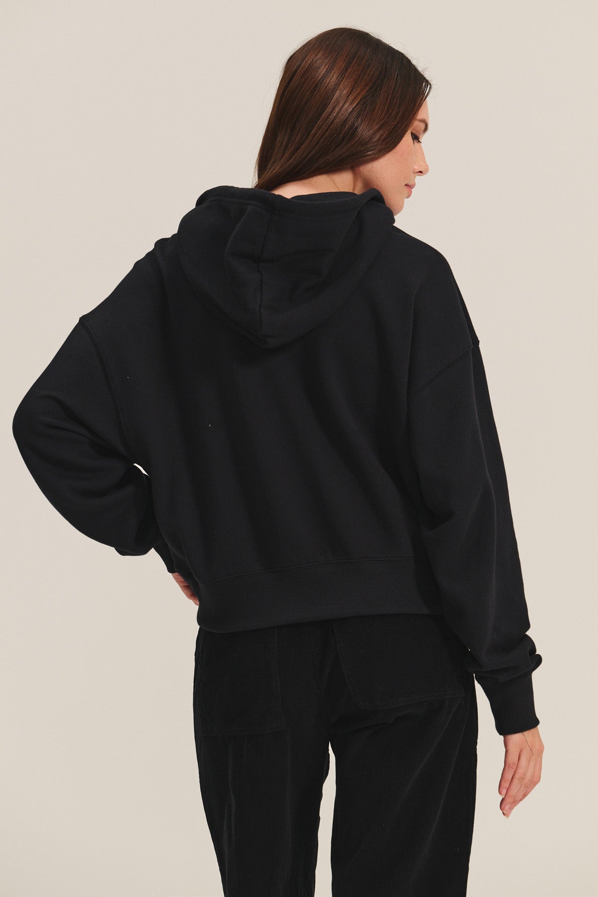 A woman with long brown hair, seen from the back against a plain light background, wears the HIGHLAND HOODIE by Velvet by Graham & Spencer paired with black pants.