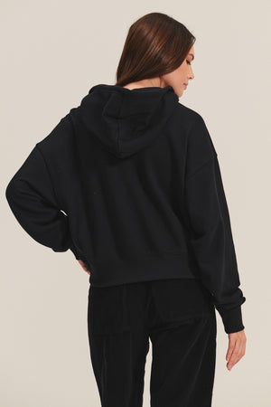 A woman with long brown hair, seen from the back against a plain light background, wears the HIGHLAND HOODIE by Velvet by Graham & Spencer paired with black pants.