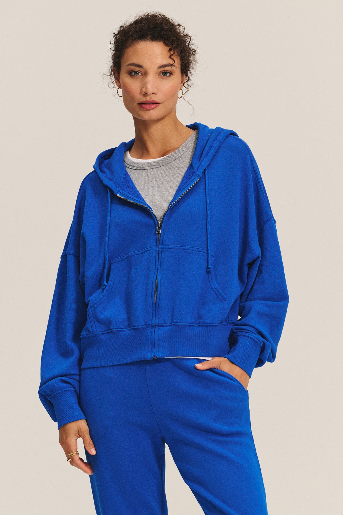 A woman models the HIGHLAND HOODIE in bright blue by Velvet by Graham & Spencer over a gray tee and matching pants, standing against a plain background and highlighting this cozy loungewear ideal for home relaxation.-40672298500289