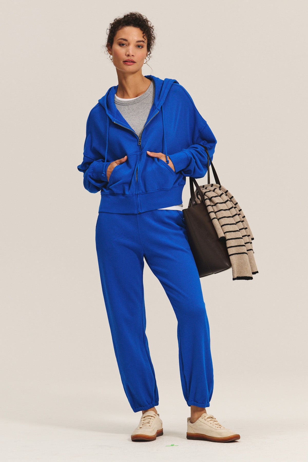 Wearing the HIGHLAND HOODIE by Velvet by Graham & Spencer in bright blue with matching joggers, this person pairs the look with a black tote and striped sweater for the ultimate comfortable loungewear style.