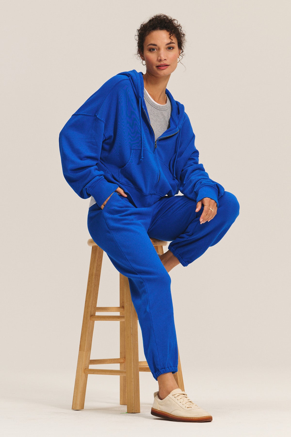 A woman sits on a wooden stool wearing the HIGHLAND HOODIE and matching sweatpants by Velvet by Graham & Spencer over a gray t-shirt, paired with beige sneakers—for ultimate cozy loungewear.-40672298434753