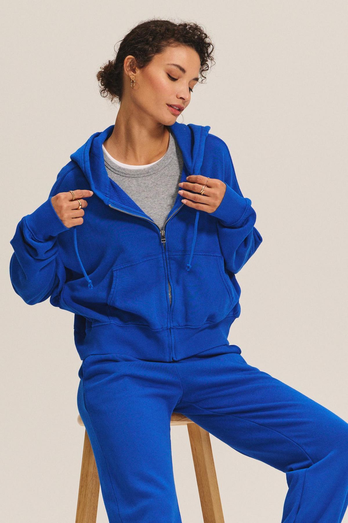 Wearing the HIGHLAND HOODIE by Velvet by Graham & Spencer over a gray tee and blue sweatpants, a person sits adjusting the strings—a perfect lounge look for relaxed days.