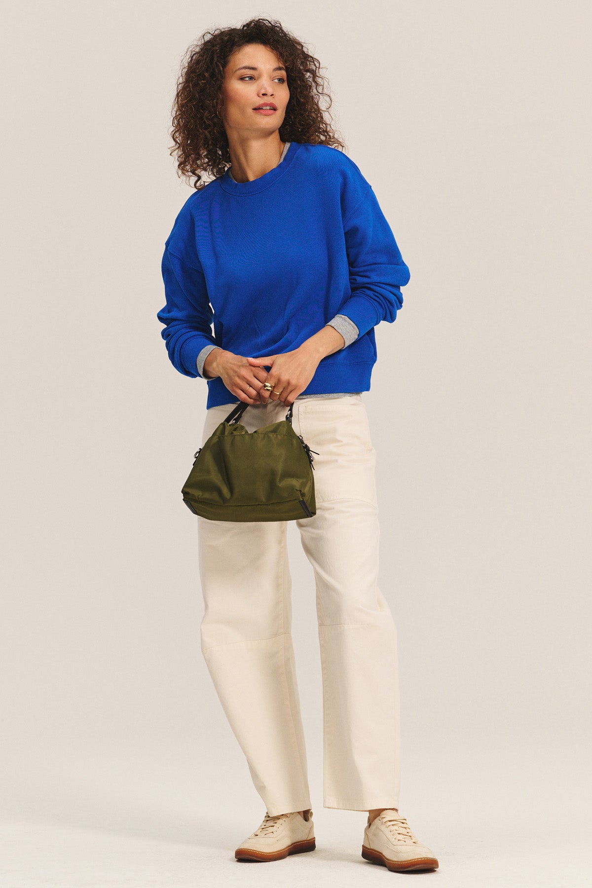 A woman wears the YOKO SWEATSHIRT by Velvet by Graham & Spencer in bright blue, paired with cream pants, beige shoes, and a green handbag, standing against a plain background.-40672504807617
