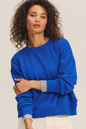A woman with curly hair wears the Velvet by Graham & Spencer YOKO SWEATSHIRT in bright blue cotton fleece over a gray long-sleeve shirt and light pants, standing against a plain background.
