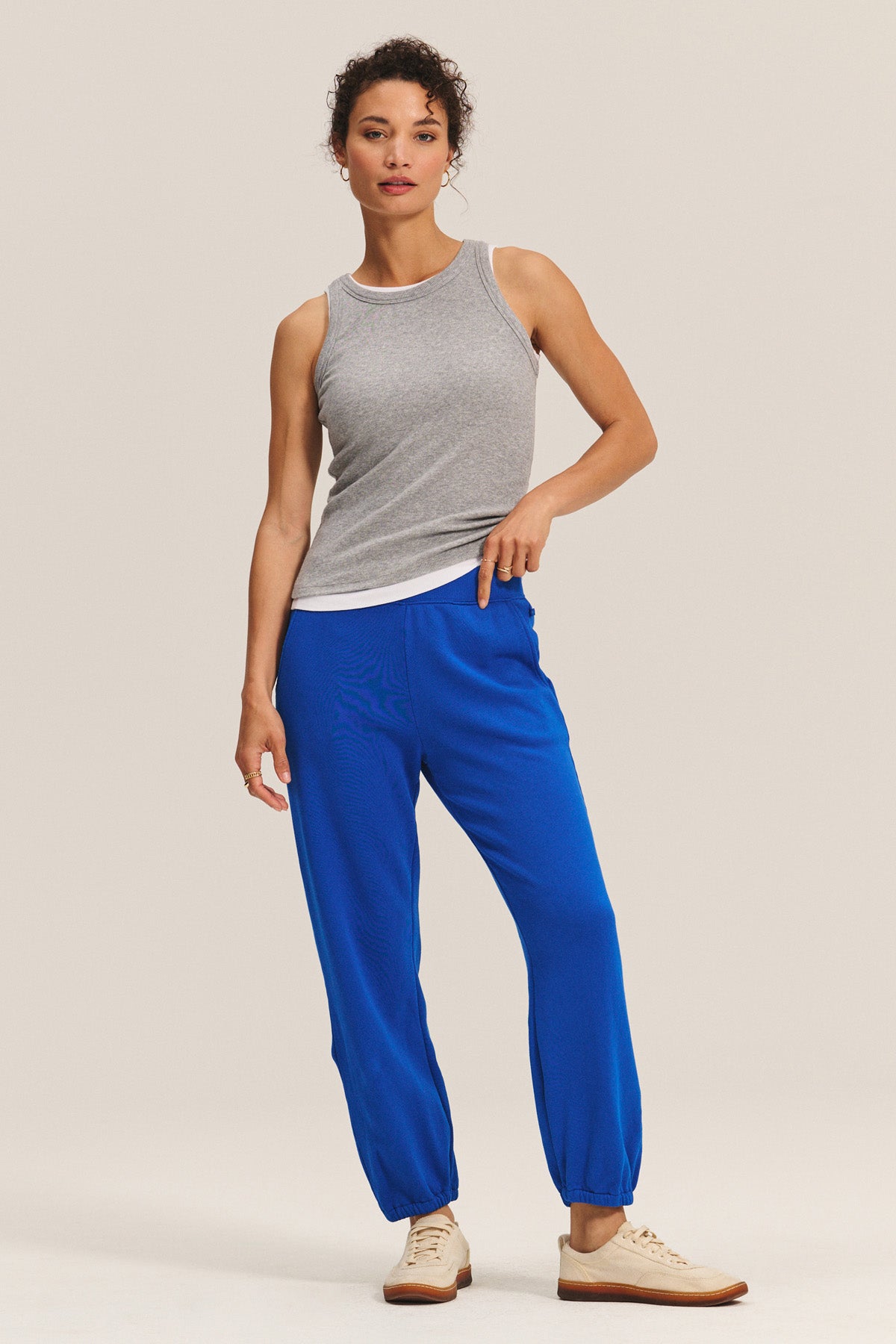 A person stands against a plain light background wearing a grey tank top, relaxed fit blue ZARA SWEATPANT by Velvet by Graham & Spencer, and beige sneakers.