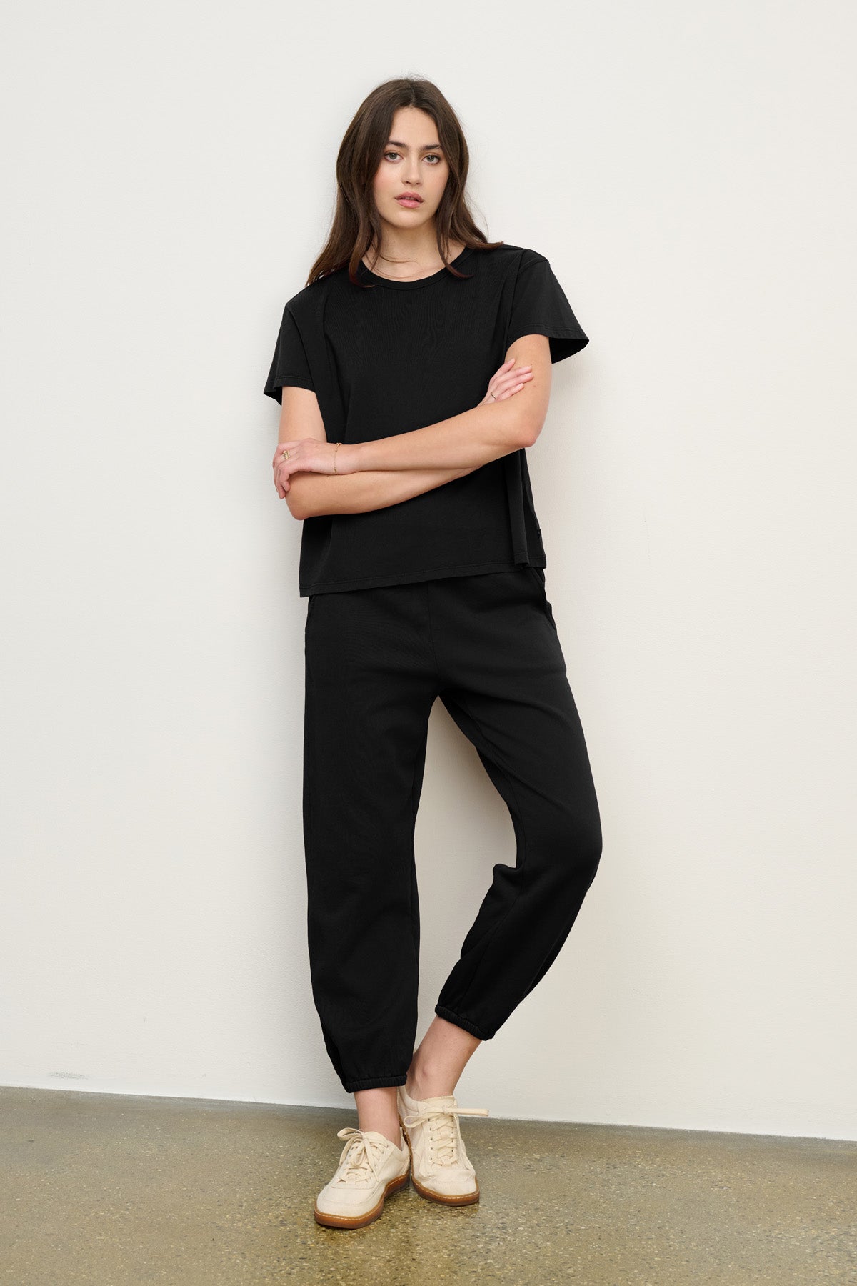 Person stands against a white wall wearing a black short-sleeve top, relaxed fit ZARA SWEATPANT by Velvet by Graham & Spencer in soft cotton fleece, and beige sneakers on a polished concrete floor.-40294545883329