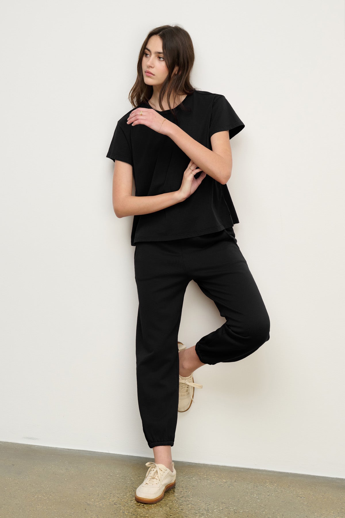 A woman stands against a white wall wearing a black short-sleeve top, Velvet by Graham & Spencer ZARA SWEATPANT in black, and beige sneakers, with one leg bent and arms crossed over her body.-40294545850561