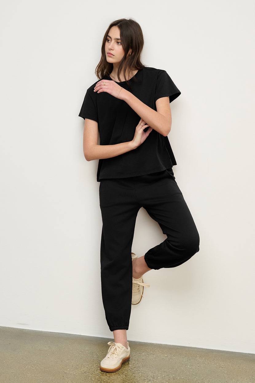 A woman poses casually against a white wall, wearing the ZUMA SWEATPANT by Velvet by Jenny Graham, a black short-sleeve top, and beige shoes.