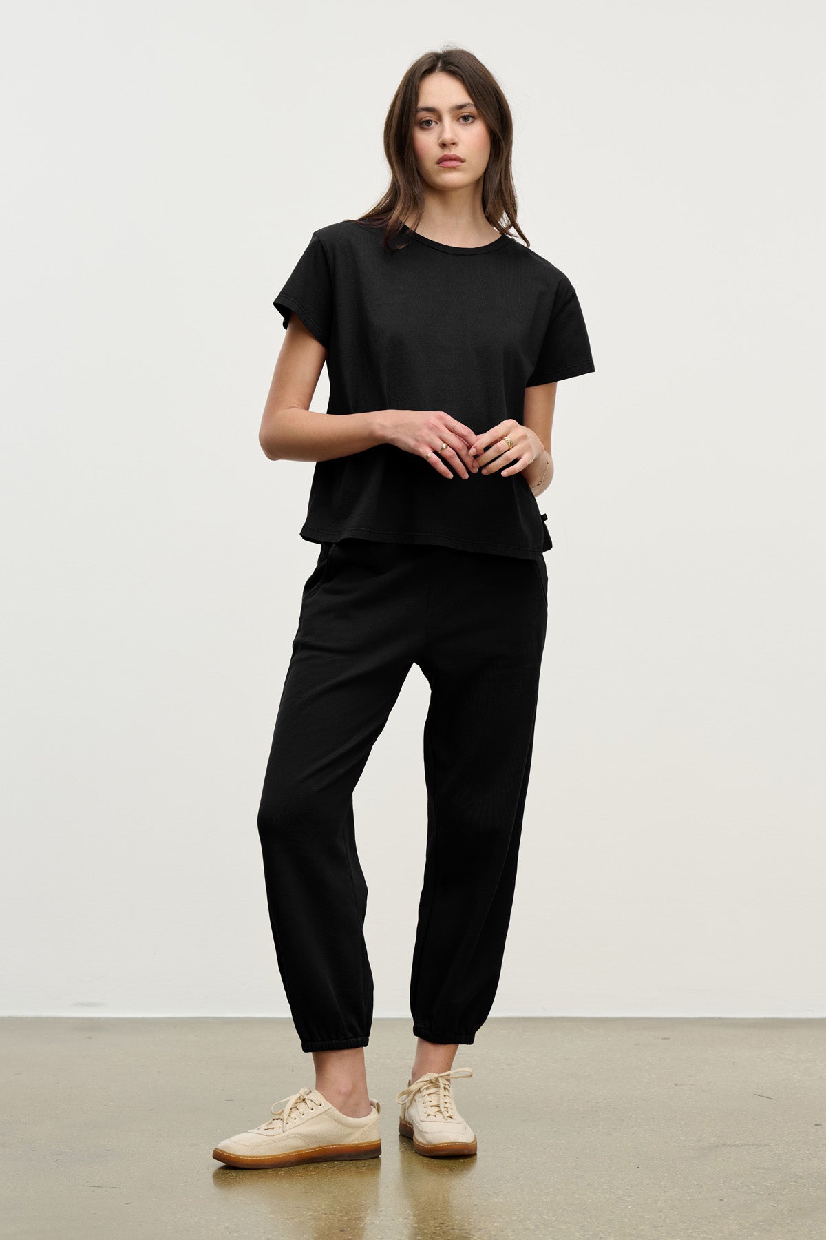 A person stands against a plain background wearing a loose black t-shirt, Velvet by Graham & Spencer ZARA SWEATPANT in black jogger style, and beige sneakers.