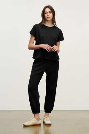 A person stands against a plain background wearing a loose black t-shirt, Velvet by Graham & Spencer ZARA SWEATPANT in black jogger style, and beige sneakers.