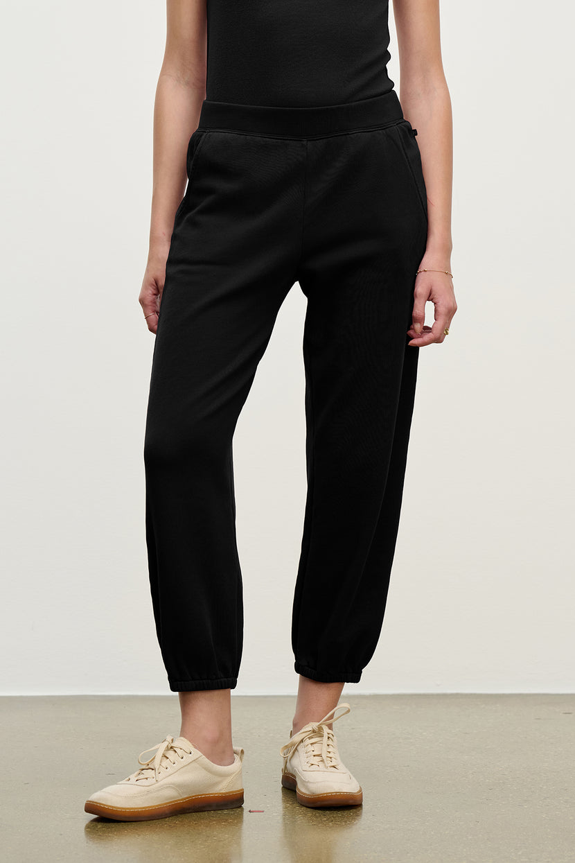 A person wearing Velvet by Jenny Graham ZUMA SWEATPANT in black organic fleece and beige sneakers stands against a plain background with hands relaxed at their sides.