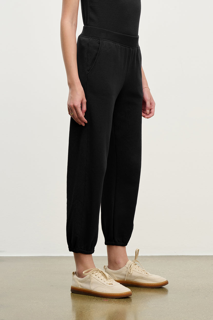 Someone wearing the ZUMA SWEATPANT in black by Velvet by Jenny Graham and beige sneakers stands against a plain white background.