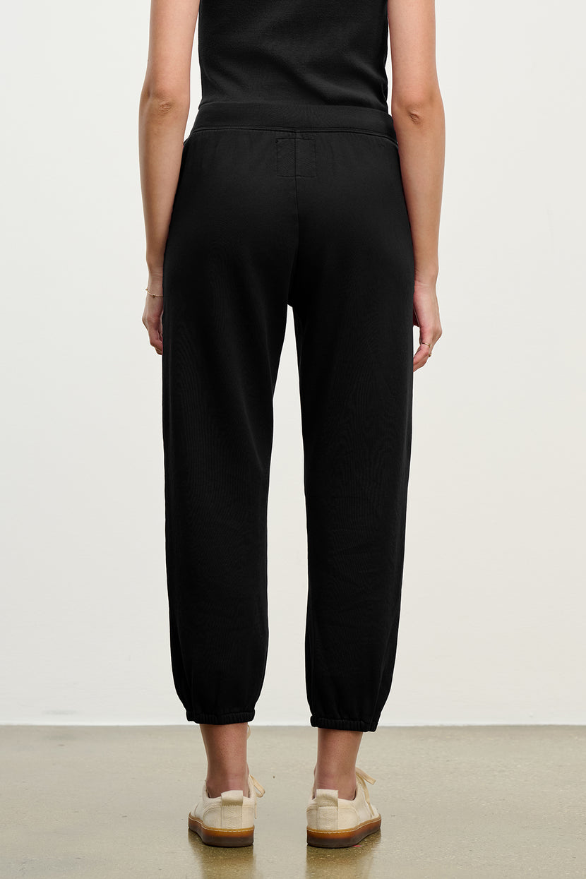 Someone is standing with their back to the camera against a plain background, wearing the ZUMA SWEATPANT by Velvet by Jenny Graham in black paired with light-colored shoes.