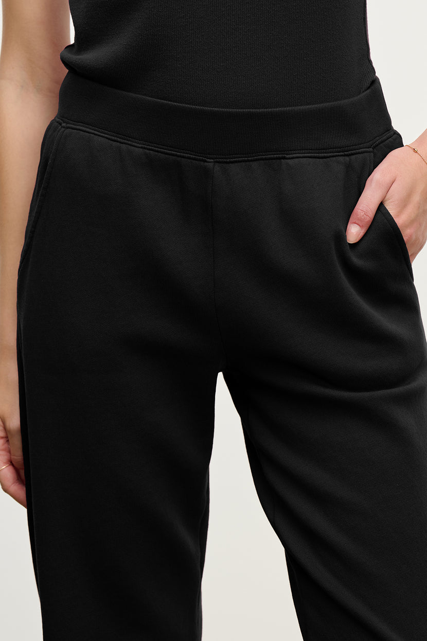 Model wearing the ZUMA SWEATPANT in black by Velvet by Jenny Graham, paired with a black top, one hand in their pocket; shown from waist to mid-thigh.